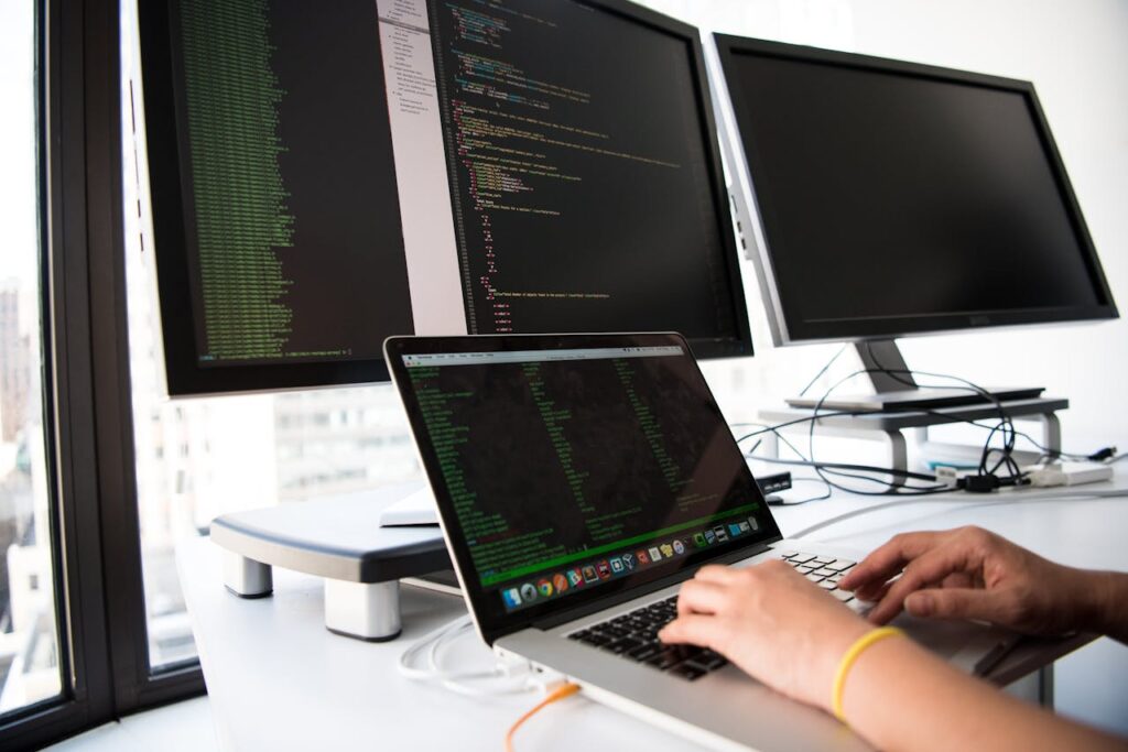pexels-photo-1181675 A developer writes code on a laptop in front of multiple monitors in an office setting.
